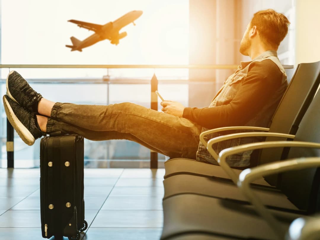A traveller waiting in an airport departure lounge, feet up on a suitcase, watching a plane take off